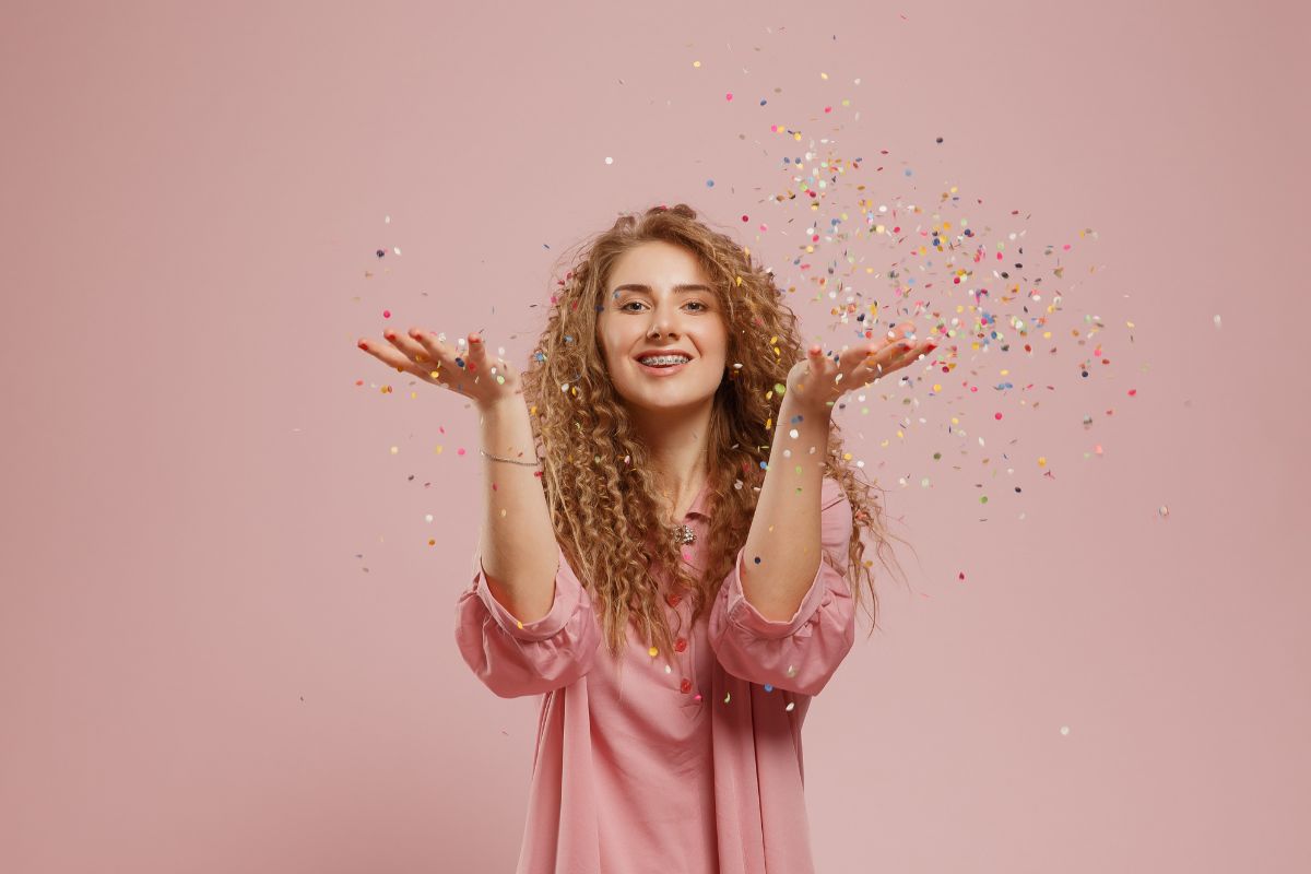 Happy woman in pink shirt throwing confetti against pink background, celebrating
